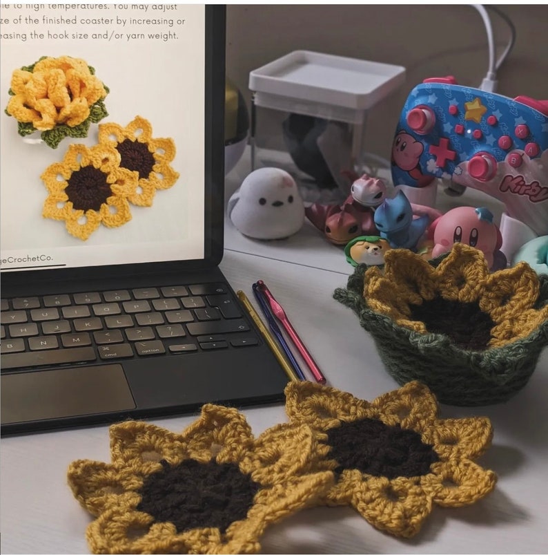Peut inclure: Deux sous-verres en forme de tournesol crochet&eacute;s en jaune et marron avec un centre vert. Les sous-verres sont sur une surface blanche &agrave; c&ocirc;t&eacute; d'un ordinateur portable, de la laine et d'aiguilles &agrave; crocheter. L'&eacute;cran de l'ordinateur portable affiche un mod&egrave;le de crochet de tournesols.