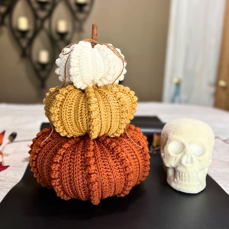 Puede incluir: Una pila de tres calabazas de ganchillo en blanco, amarillo mostaza y naranja quemado, cada una con un dise&ntilde;o acanalado texturizado y un peque&ntilde;o tallo marr&oacute;n en la parte superior. Junto a las calabazas hay una r&eacute;plica de cr&aacute;neo humano blanquecino.