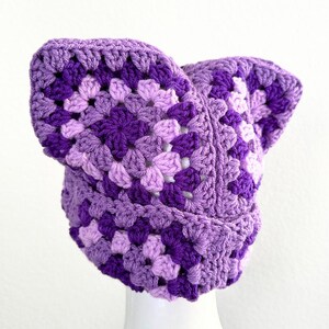May include: A purple and lavender crocheted hat with a unique ear flap design.