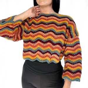 May include: A long-sleeved crochet sweater featuring a horizontal chevron pattern in alternating stripes of mustard yellow, burnt orange, deep burgundy, and muted slate blue. The garment has a wide neckline and a cropped, loose-fitting silhouette.