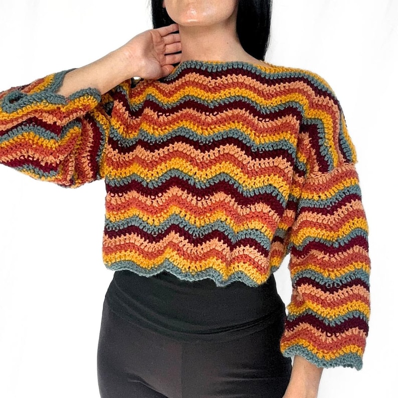 May include: A long-sleeved crochet sweater featuring a horizontal chevron pattern in alternating stripes of mustard yellow, burnt orange, deep burgundy, and muted slate blue. The garment has a wide neckline and a cropped, loose-fitting silhouette.
