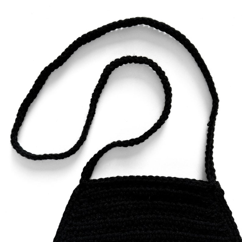 May include: A black crocheted bag featuring a textured, hand-stitched yarn construction. The accessory includes a long, thin, matching black crocheted strap that is looped and attached to the top edge of the rectangular-shaped bag body.