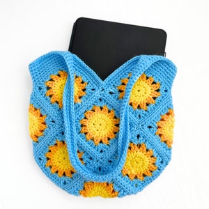 May include: A handmade crochet tote bag constructed from blue square motifs featuring yellow and orange sunburst patterns. The bag has two integrated blue handles and contains a rectangular black electronic device partially visible inside the opening.