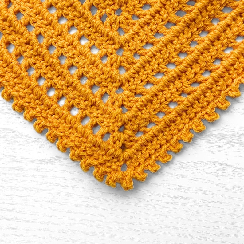 Puede incluir: Primer plano de un cuadrado amarillo tejido a crochet con un patr&oacute;n de encaje. El cuadrado est&aacute; sobre una superficie de madera blanca.