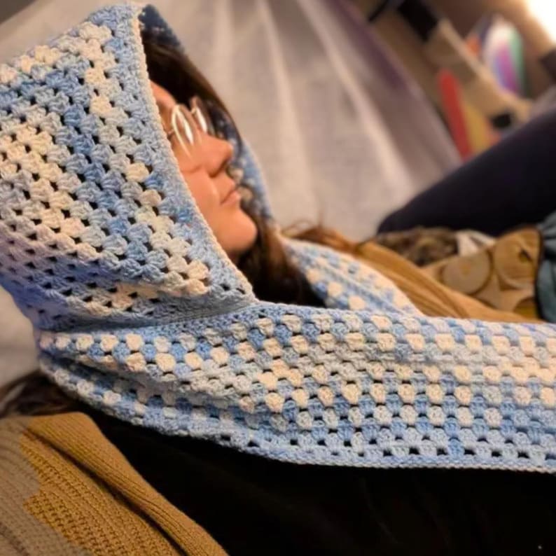 May include: A hooded scarf crafted with a crochet granny square pattern in alternating shades of light blue and white. The garment features a connected hood and a long scarf section, showcasing a consistent open-work stitch throughout the design.