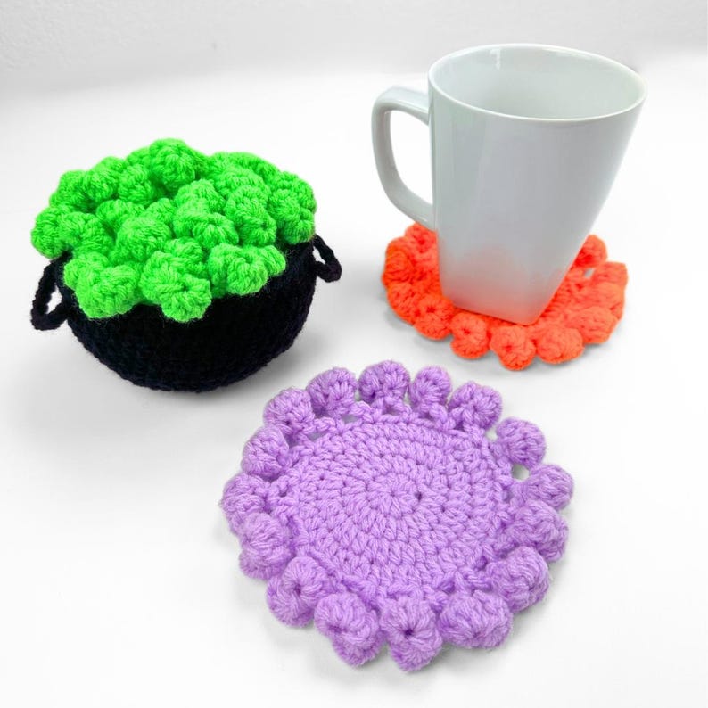 May include: A black crocheted cauldron filled with bright green crocheted bubbles, a white ceramic mug resting on an orange crocheted coaster, and a separate purple crocheted coaster with a textured, scalloped edge design.