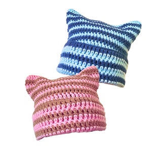 Op de afbeelding: Twee gebreide mutsen met kattenoren. De ene muts is roze en bruin gestreept, de andere is blauw en lichtblauw gestreept.