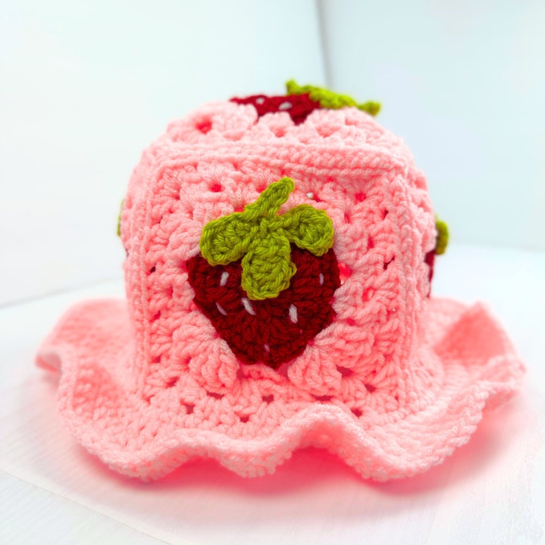 K&ouml;nnte beinhalten: Ein rosa geh&auml;kelter Sonnenhut mit Erdbeer-Applikationen. Der Hut hat eine breite Krempe und einen gekr&auml;uselten Rand.