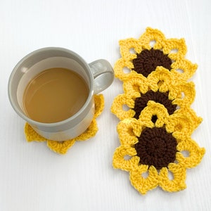 Peut inclure: Un ensemble de quatre dessous-de-verre en crochet en forme de tournesol, jaunes et bruns. Les dessous-de-verre sont empil&eacute;s les uns sur les autres. Une tasse de caf&eacute; repose sur un dessous-de-verre.