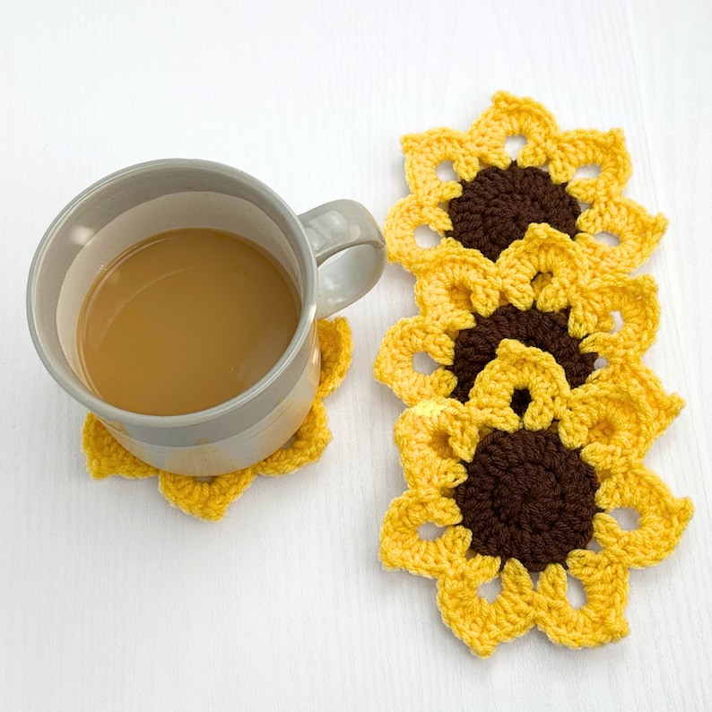 Peut inclure: Un ensemble de quatre dessous-de-verre en crochet en forme de tournesol, jaunes et bruns. Les dessous-de-verre sont empil&eacute;s les uns sur les autres. Une tasse de caf&eacute; repose sur un dessous-de-verre.