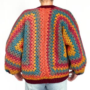 May include: A colorful crochet cardigan with a geometric pattern in shades of orange, red, blue, brown, and yellow. The cardigan has a V-neck and long sleeves.