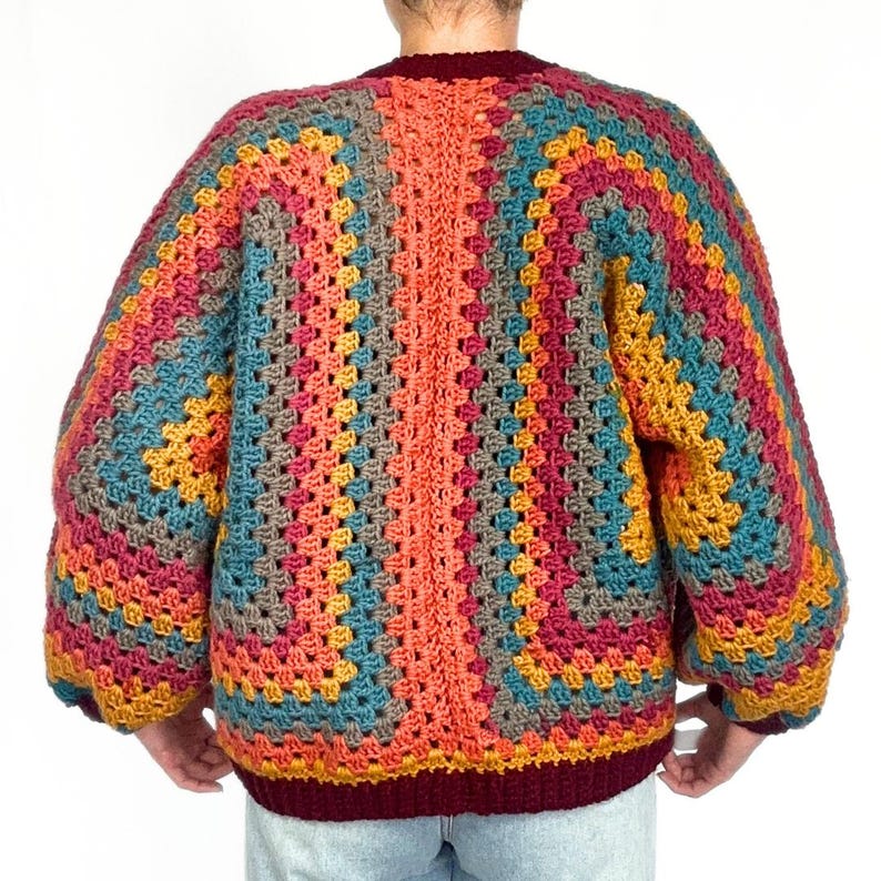 May include: A colorful crochet cardigan with a geometric pattern in shades of orange, red, blue, brown, and yellow. The cardigan has a V-neck and long sleeves.