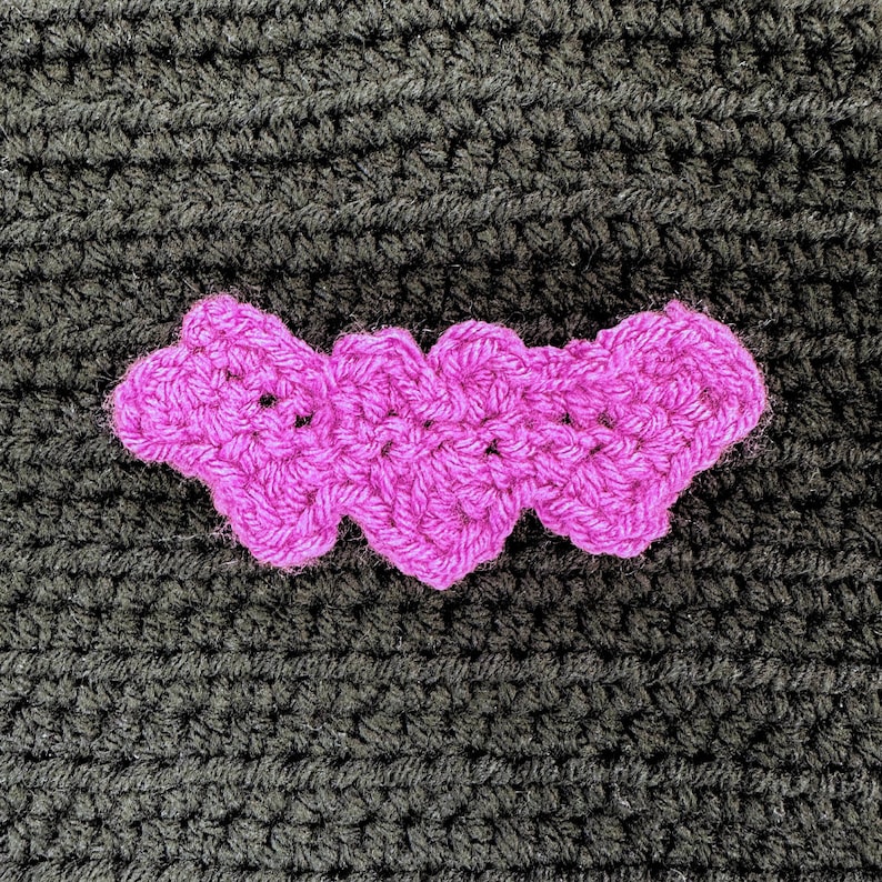 May include: A bright magenta crochet applique shaped like a bat with scalloped wing edges. The piece is crafted from yarn with a visible looped texture and is positioned against a dark black crocheted background with a ribbed horizontal pattern.