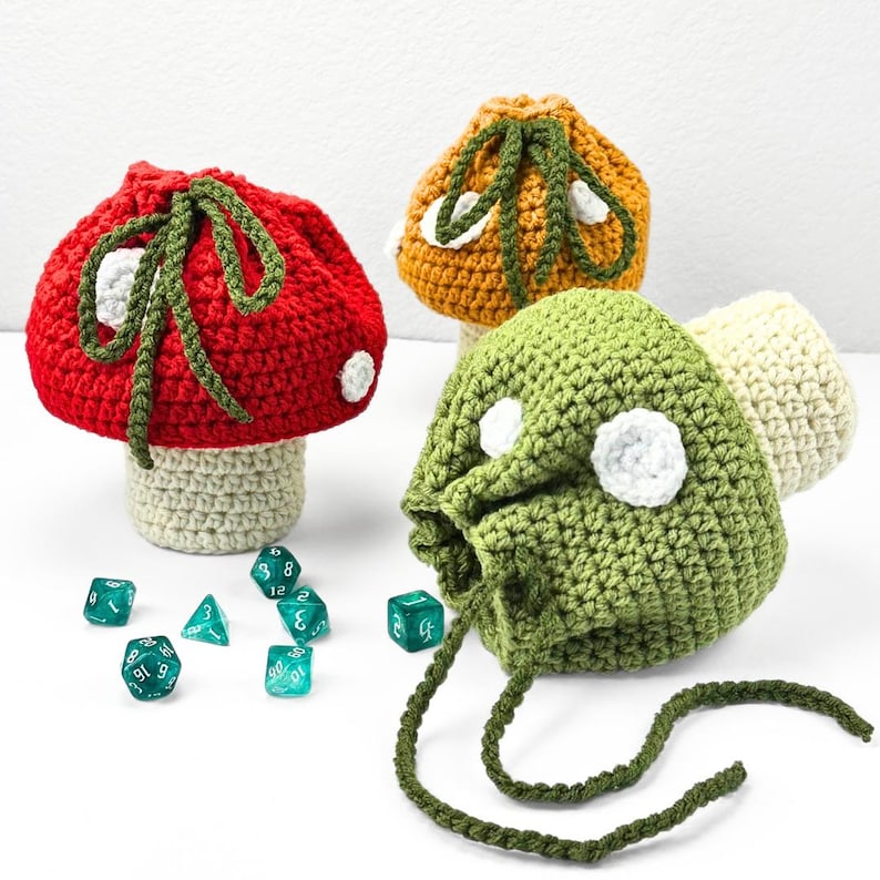 May include: Three crochet mushroom-shaped drawstring pouches in red, orange, and green, each featuring white spots and a beige stem base. The pouches have dark green crocheted drawstrings. Several translucent teal polyhedral dice are scattered nearby.