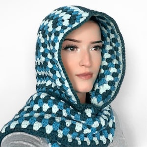 May include: A hooded cowl crafted with a crochet granny square pattern in shades of dark teal, medium blue, and pale light blue. The hood features a thick, solid dark teal border along the front edge, framing the face with a textured, yarn-based design.