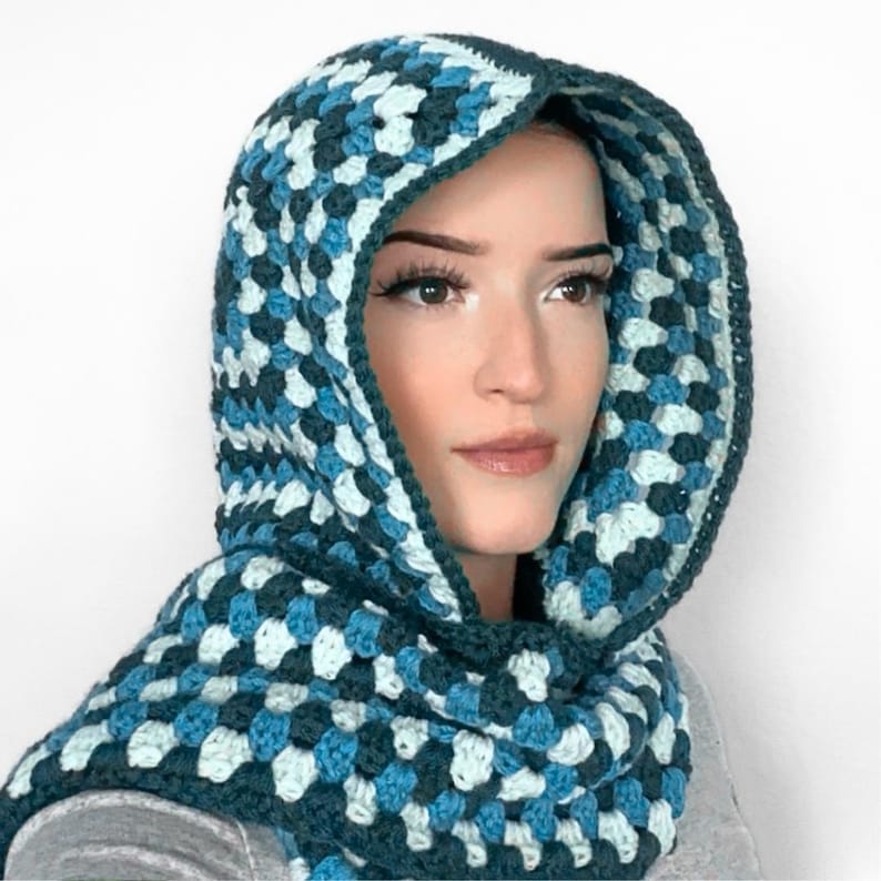 May include: A hooded cowl crafted with a crochet granny square pattern in shades of dark teal, medium blue, and pale light blue. The hood features a thick, solid dark teal border along the front edge, framing the face with a textured, yarn-based design.