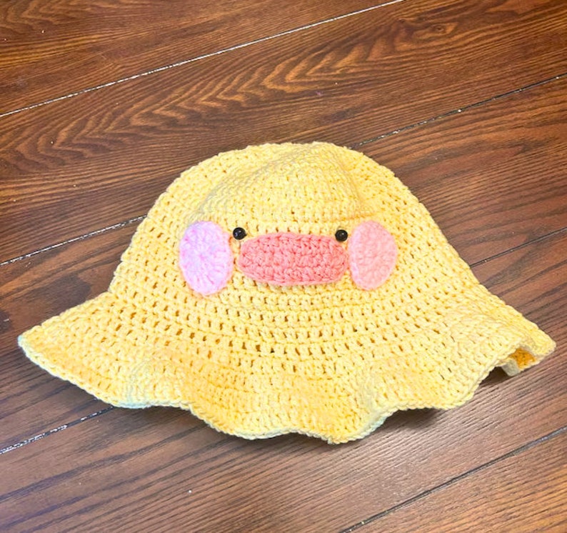 Pu&ograve; includere: Un cappello a secchiello di uncinetto giallo con un design a forma di faccia di anatra rosa e nera.