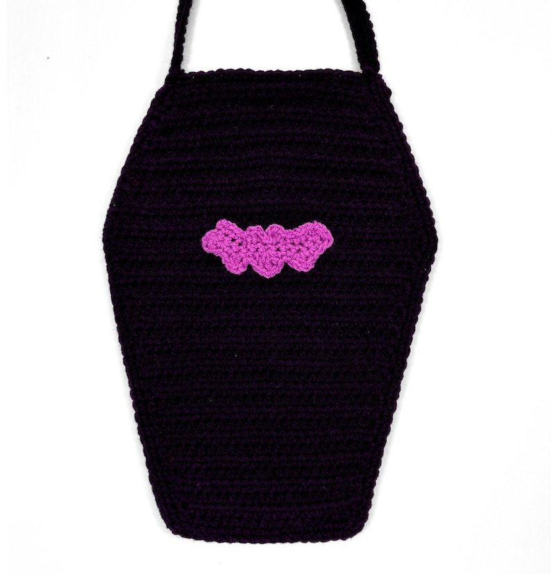 May include: A black crocheted bag shaped like a coffin, featuring a bright pink crocheted bat appliqu&eacute; centered on the front. The bag includes a black crocheted strap attached to the top, showcasing a textured yarn construction throughout the piece.