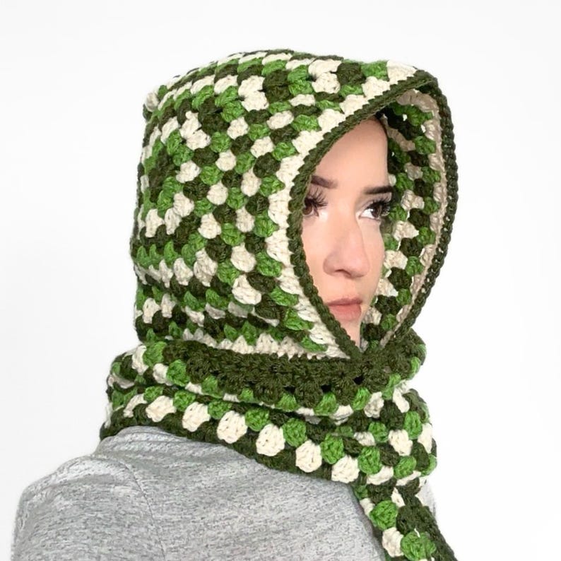 May include: A crochet hood and scarf combination featuring a granny square pattern in shades of dark green, light green, and cream. The hood is designed to frame the face, while the attached scarf drapes down, showcasing a consistent yarn texture.