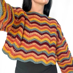 May include: A long-sleeved crochet sweater featuring a horizontal chevron wave pattern in alternating colors of mustard yellow, burnt orange, deep burgundy, and muted slate blue. The garment has a round neckline and a cropped, scalloped hemline.