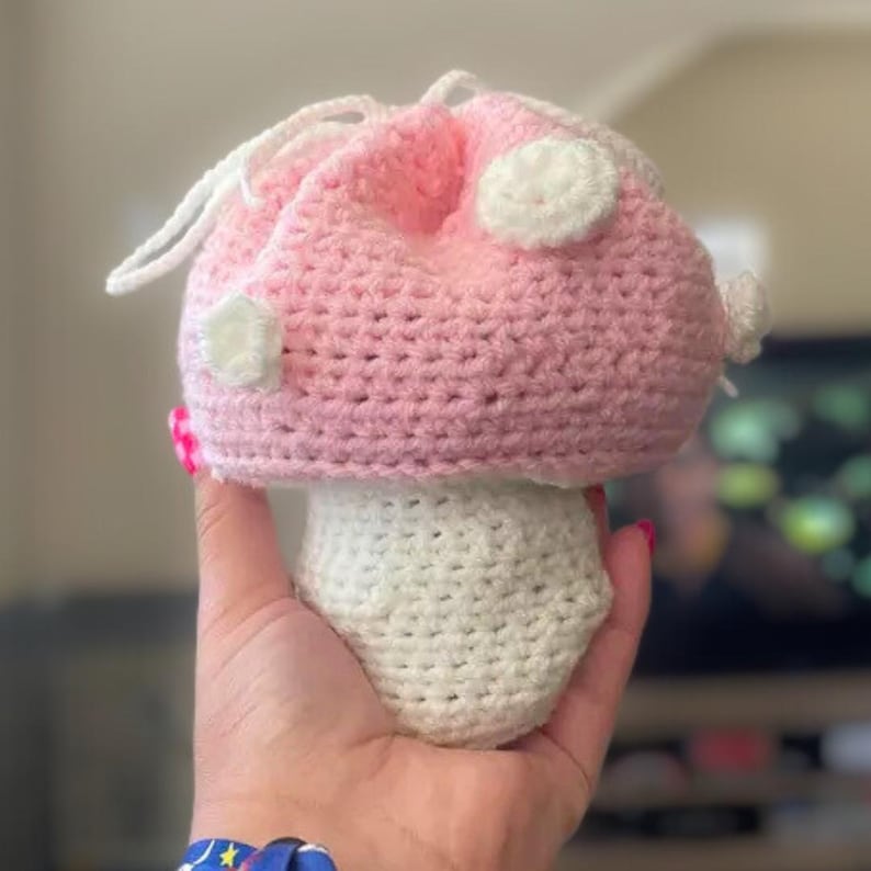 May include: A crochet drawstring pouch shaped like a mushroom with a light pink cap featuring white circular spots and a white stem. The top of the cap includes white yarn strings for closing the pouch, creating a soft, textured appearance.