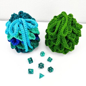 May include: Two crocheted drawstring pouches with a textured scale-like pattern. One pouch features a gradient of blue and green yarn with light blue ties, while the other is solid green. Seven translucent green polyhedral dice are arranged below.