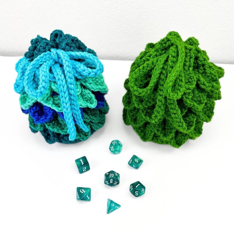 May include: Two crocheted drawstring pouches with a textured scale-like pattern. One pouch features a gradient of blue and green yarn with light blue ties, while the other is solid green. Seven translucent green polyhedral dice are arranged below.