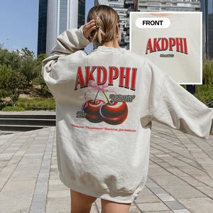 May include: Oversized beige sweatshirt with the text "AKDPHI" in red on the front and back. The back features a graphic of two cherries with the text "The Sweetest Weekend" and "Since 2023".