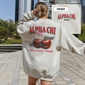May include: A cream colored sweatshirt with the text "Alpha Chi" in red letters and a graphic of two cherries with the text "The Sweetest Sorority" and "Real Strong Women".