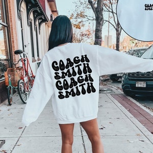 Könnte beinhalten: Weißer Pullover mit dem Text "COACH SMITH", der zweimal in einer Retro-Schrift auf der Rückseite wiederholt wird.