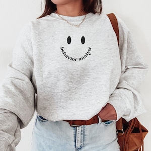 Op de afbeelding: Grijs sweatshirt met een smiley-gezicht en de tekst "behavior analyst" eronder gedrukt.
