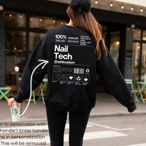 Op de afbeelding: Zwart sweatshirt met witte tekst "Nail Tech @addcustom" en een afbeelding op de achterkant. Het sweatshirt heeft ook extra tekst en pictogrammen. De persoon draagt een zwarte legging en een zwarte pet.