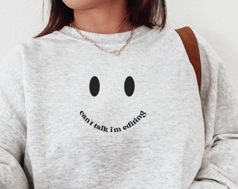 May include: A light gray sweatshirt with a black smiley face design and the text "can't talk I'm editing". The sweatshirt has long sleeves and a crew neck. The model is wearing light blue jeans and a brown belt.