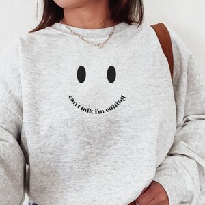 May include: A light gray sweatshirt with a black smiley face design and the text "can't talk I'm editing". The sweatshirt has long sleeves and a crew neck. The model is wearing light blue jeans and a brown belt.