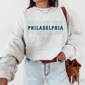Puede incluir: Una sudadera gris claro con la palabra "PHILADELPHIA" impresa en verde azulado y blanco. El texto est&aacute; apilado verticalmente, con el nombre de la ciudad en negrita. La sudadera tiene mangas largas.