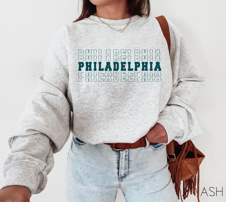 Puede incluir: Una sudadera gris claro con la palabra "PHILADELPHIA" impresa en verde azulado y blanco. El texto est&aacute; apilado verticalmente, con el nombre de la ciudad en negrita. La sudadera tiene mangas largas.