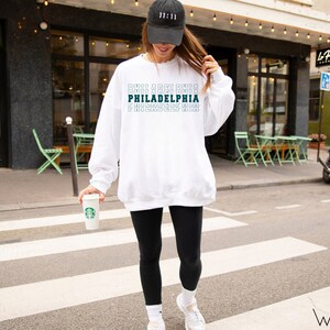 Puede incluir: Sudadera blanca con la palabra "PHILADELPHIA" en un dise&ntilde;o apilado. El texto es de color verde azulado. La persona de la imagen lleva una gorra de b&eacute;isbol negra con el n&uacute;mero "11:11", leggings negros y zapatillas blancas.