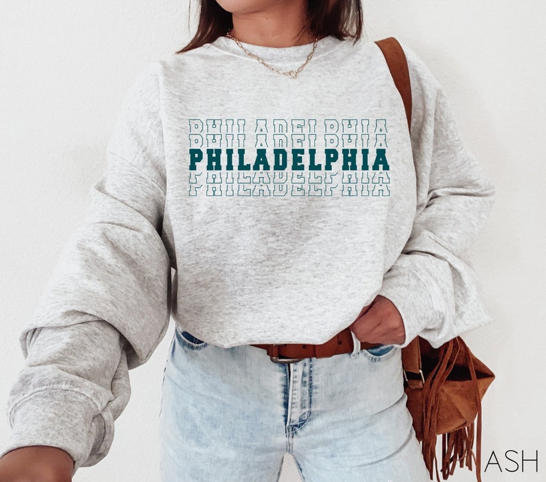 Puede incluir: Una sudadera gris claro con la palabra "PHILADELPHIA" en letras de bloque verde azulado y blanco. La palabra est&aacute; apilada varias veces. La sudadera tiene mangas largas y se combina con vaqueros de lavado claro y un cintur&oacute;n marr&oacute;n.