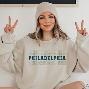 Puede incluir: Sudadera de color arena con la palabra "PHILADELPHIA" en un dise&ntilde;o apilado. El texto es de color verde azulado y blanco, con una fuente retro. La sudadera se combina con un gorro a juego.