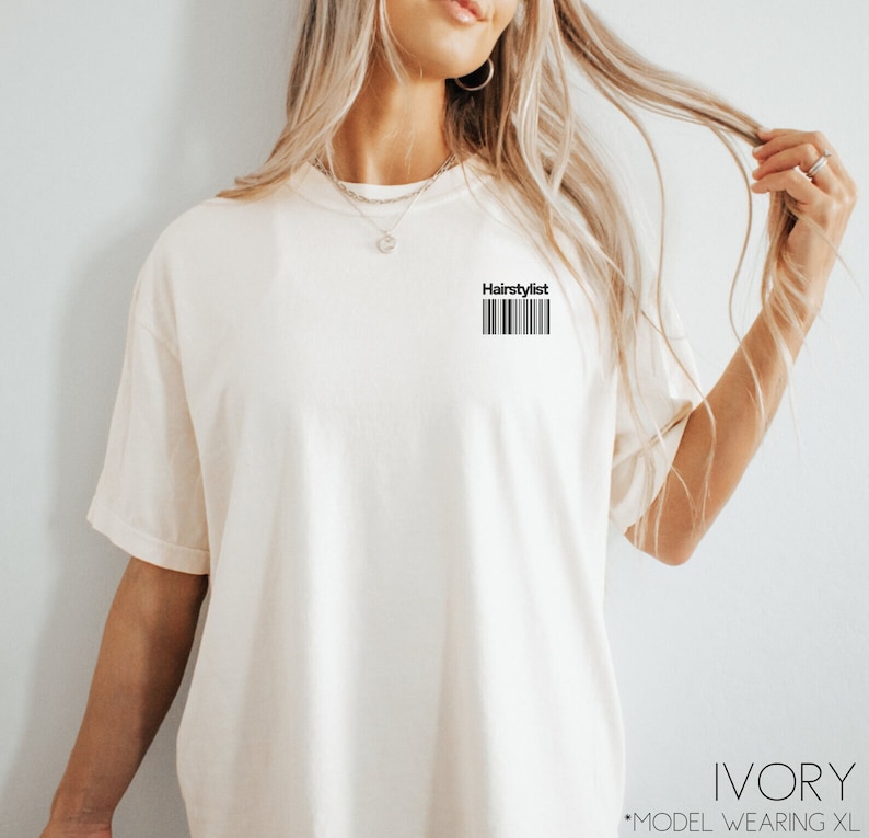 May include: Ivory t-shirt with the text "Hairstylist" and a barcode printed on the front. The model is wearing a size XL.