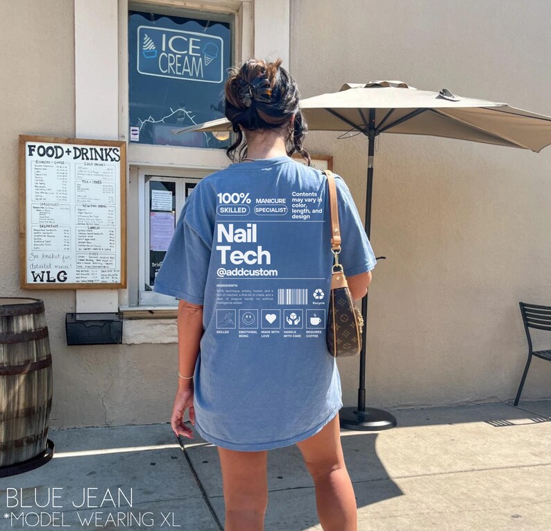 Pu&ograve; includere: Una t-shirt blu jeans con testo bianco che recita "100% Skilled Manicure Specialist Nail Tech @addcustom". Il testo &egrave; in un riquadro con un codice a barre e altri elementi grafici. La t-shirt &egrave; indossata da una persona in piedi davanti a un edificio.