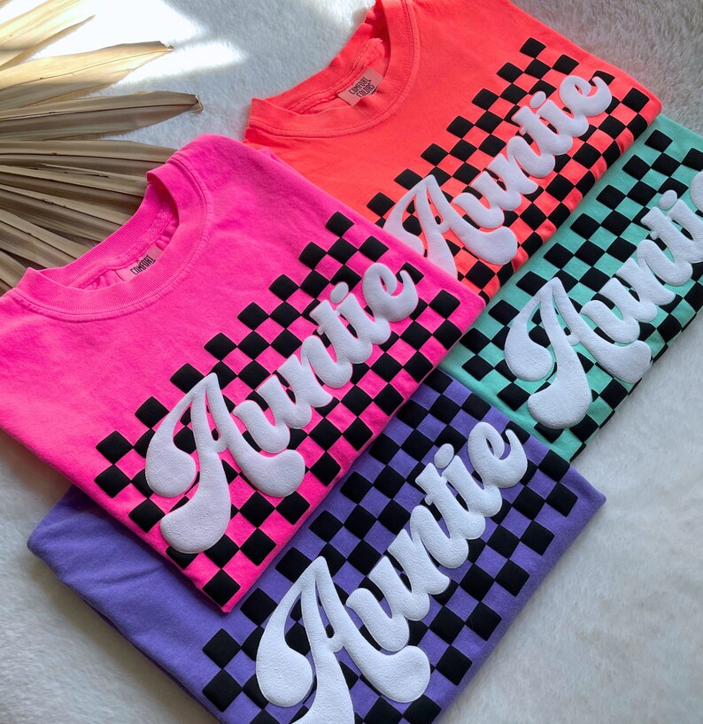 May include: Four t-shirts with a checkered pattern and the word "Auntie" in a retro font. The shirts are in pink, orange, green, and purple.