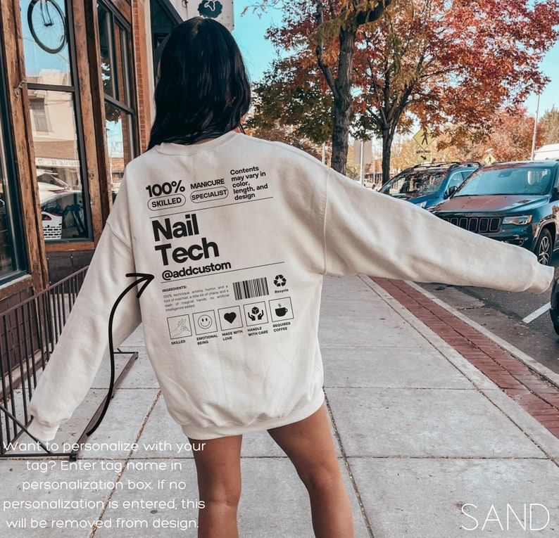 May include: A sand-colored sweatshirt with the text "Nail Tech" and "@addcustom" printed on the back. The design includes additional text and icons, such as "100% Skilled Manicure Specialist." The sweatshirt is a casual garment.