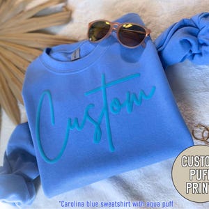 May include: A light blue sweatshirt with a turquoise "Custom" puff print. The sweatshirt is folded and laid on a white fuzzy surface. Sunglasses and earrings are also visible in the image.