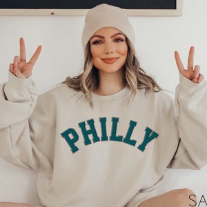 May include: A woman wearing a beige sweatshirt with the word "PHILLY" in teal green letters. She is wearing a beige beanie and has her hands up in a peace sign.