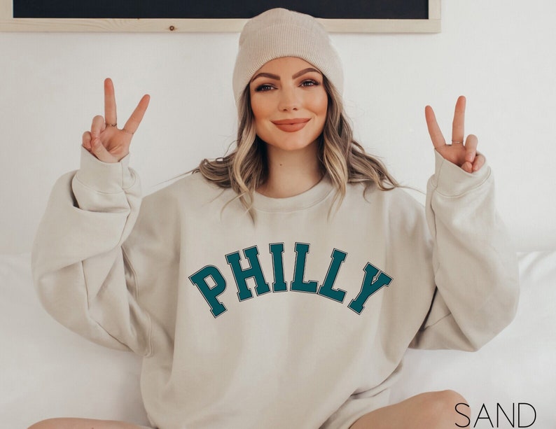 May include: A woman wearing a beige sweatshirt with the word "PHILLY" in teal green letters. She is wearing a beige beanie and has her hands up in a peace sign.