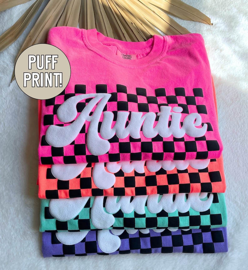 May include: Stack of four t-shirts with a checkered pattern and the word "Auntie" in a white puffy font. The shirts are pink, orange, green, and purple.
