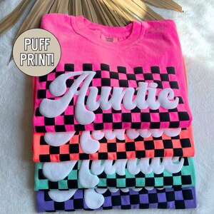 May include: A stack of four t-shirts with a checkered pattern and the word "Auntie" in a puffy white font. The shirts are in pink, orange, green, and purple.