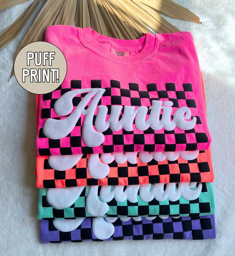 May include: A stack of four t-shirts with a checkered pattern and the word "Auntie" in a puffy white font. The shirts are in pink, orange, green, and purple.