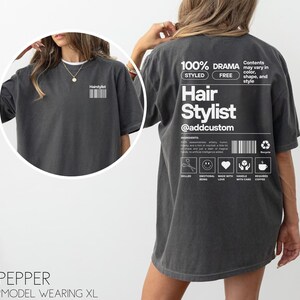 May include: A dark grey t-shirt with the text "Hair Stylist @addcustom" printed on the back. The shirt also features a graphic of a pair of scissors, a smiley face, a heart, a hand with care symbol, and a coffee cup.